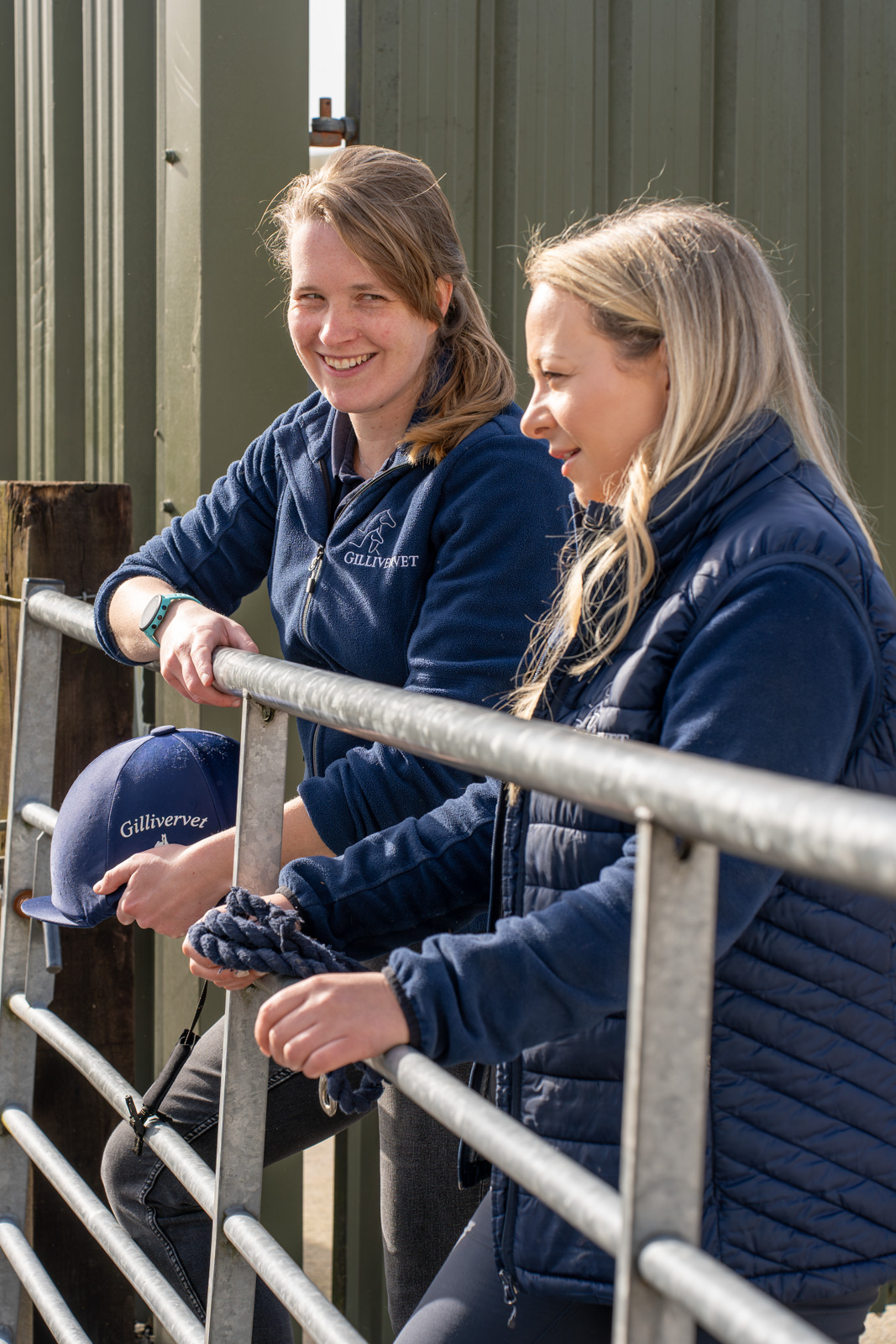Two women in navy blue jackets are standing by a railing outside. One is holding a blue cap with Gilliver on it, while the other holds a rope. They are smiling and engaged in conversation, with a green structure in the background.