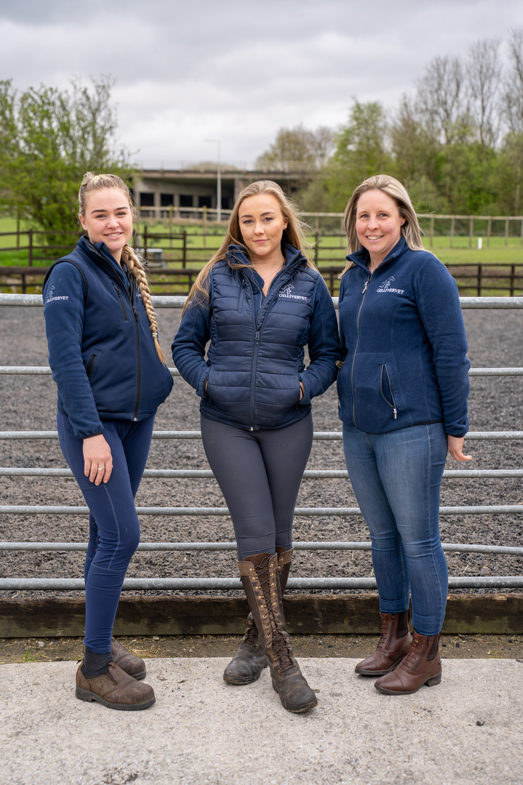 Three women stand outdoors on a gravel surface, wearing blue zip-up jackets and boots. Trees and a fence are visible in the background under an overcast sky.